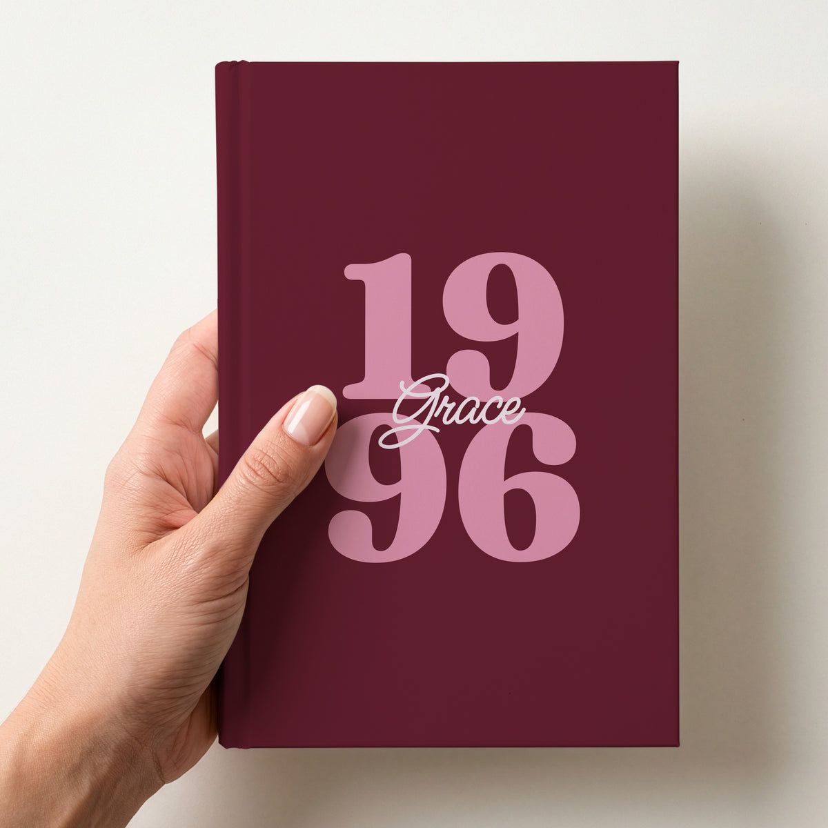 Hand holding a burgundy personalised journal with pink 1996 birth-year numbers and the name Grace