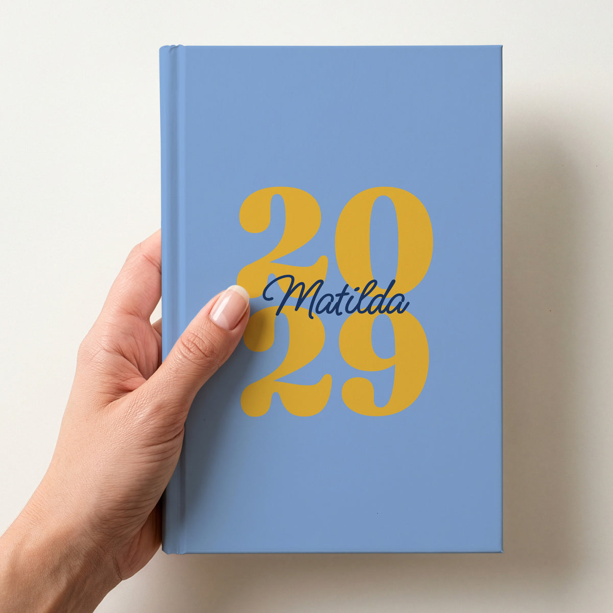 Hand holding a sky blue personalised journal with yellow 2029 numbers and the name Matilda in navy script