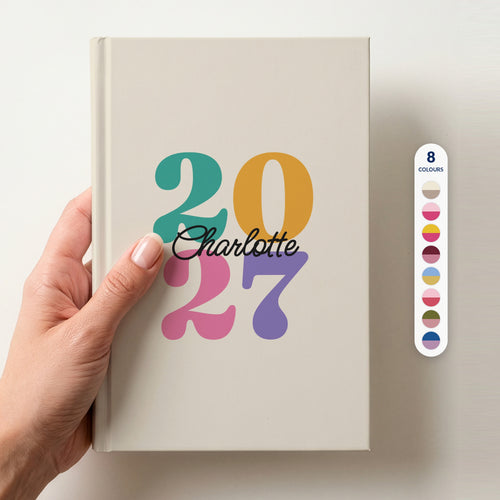 Hand holding an ivory personalised journal with rainbow 2027 numbers and the name Charlotte in script, beside a column of 8 colour swatches showing all 8 colourways