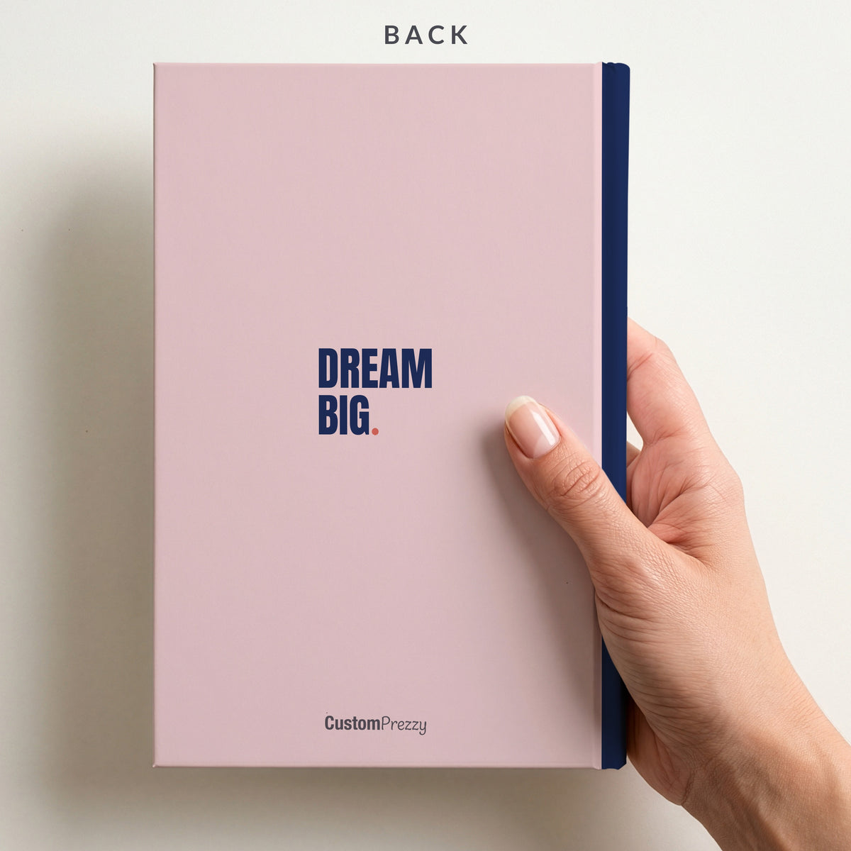 Back of the blush and navy notebook held in a hand, with the Custom Prezzy wordmark at the foot