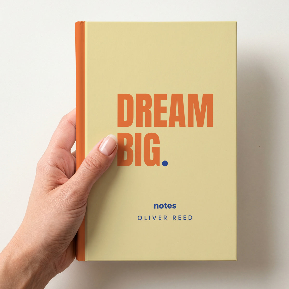 Hand holding a butter personalised notebook with 'DREAM BIG.' in tangerine and 'notes' and the name Oliver Reed below