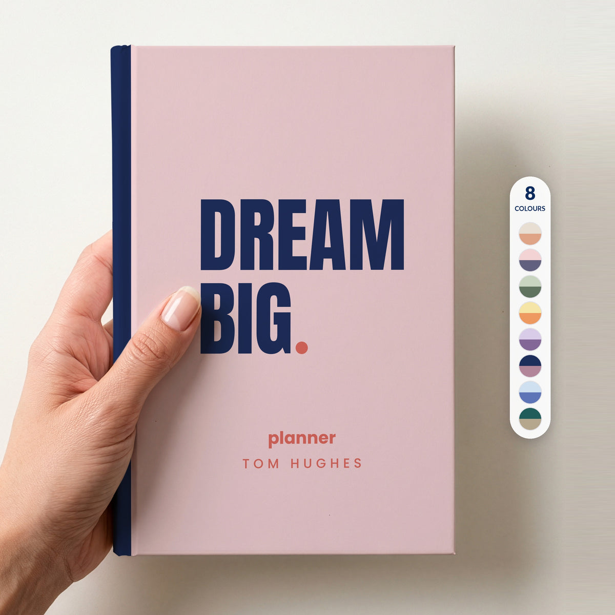 Hand holding a blush personalised notebook with 'DREAM BIG.' in navy and 'planner' and the name Tom Hughes below, beside a column of 8 colour swatches showing all 8 colourways