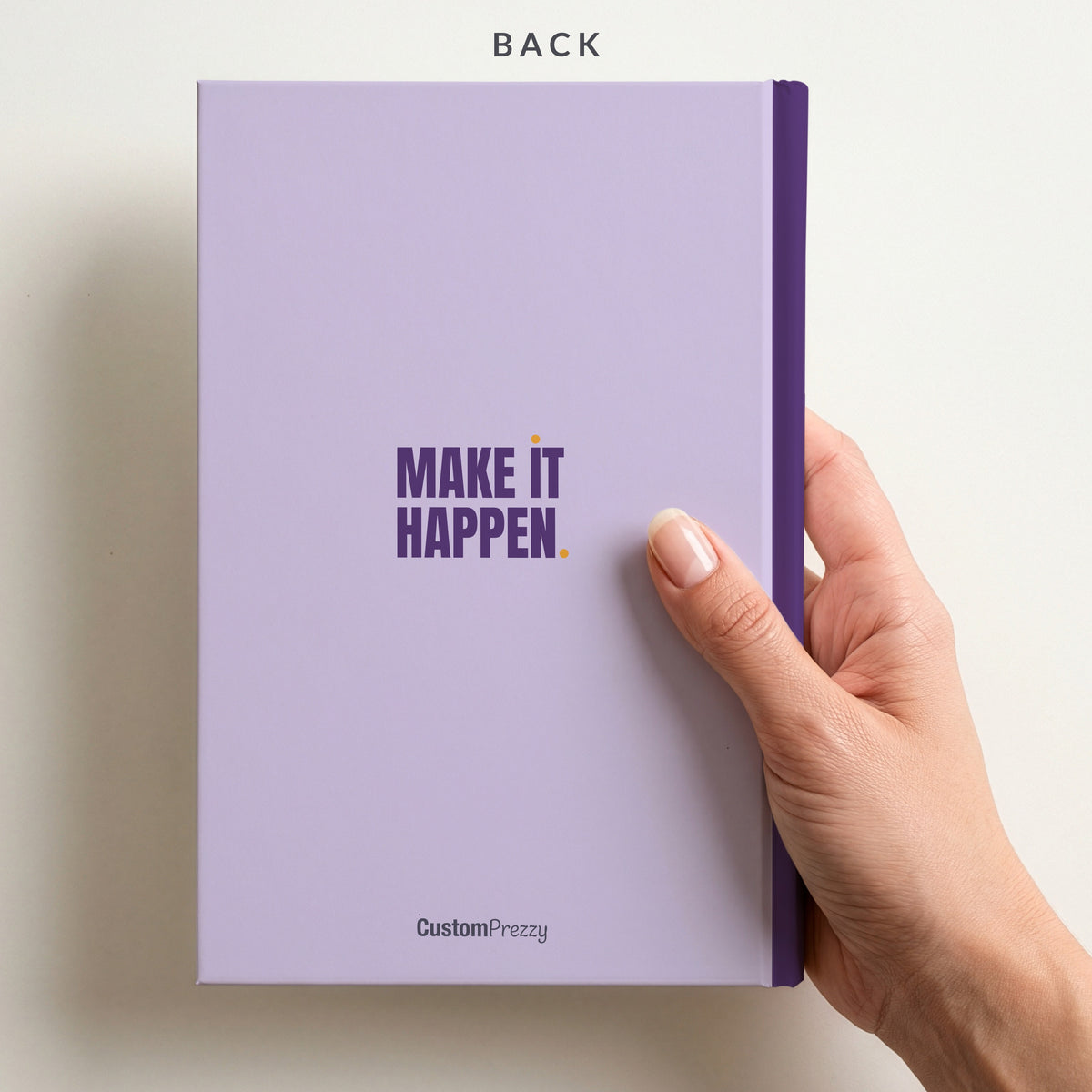 Back of the lilac & plum 'MAKE IT HAPPEN.' notebook held in a hand, with a small slogan, a contrast spine and the Custom Prezzy wordmark at the foot