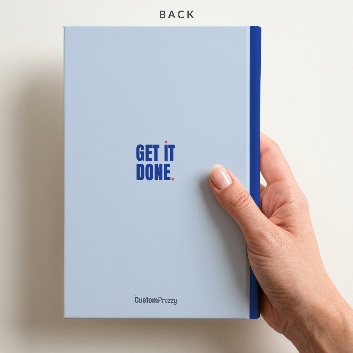 Back of the sky & cobalt 'GET IT DONE.' notebook held in a hand, with a small slogan, a contrast spine and the Custom Prezzy wordmark at the foot