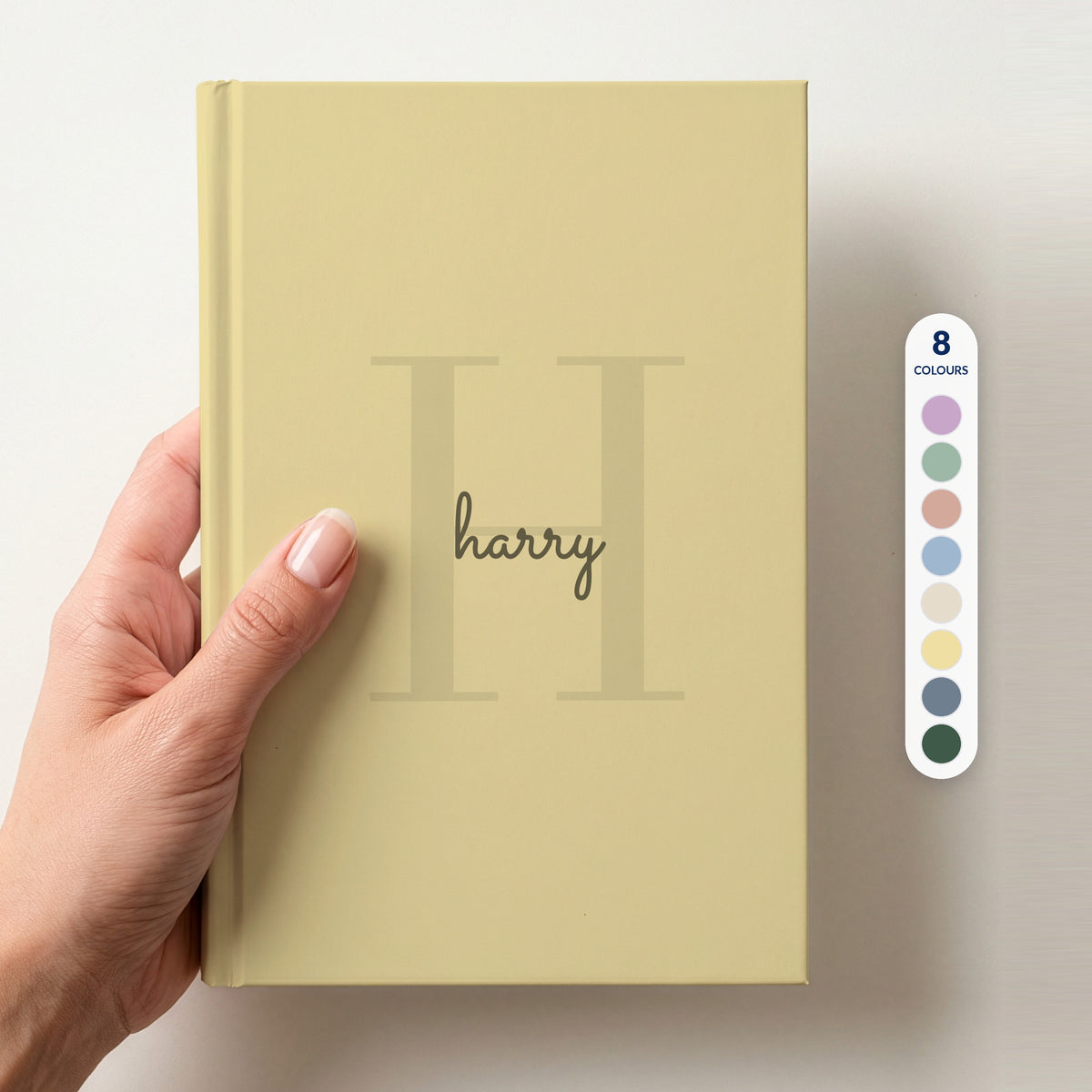 Hand holding a butter personalised journal with a large H initial and the name harry in script, beside a column of 8 colour swatches showing all 8 colourways