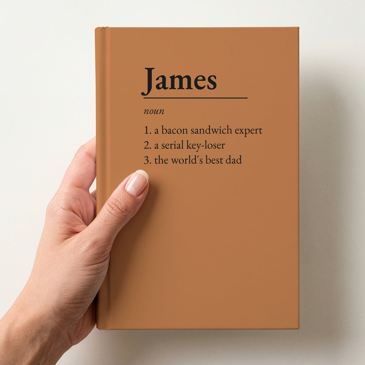 Hand holding a caramel personalised notebook with a dictionary definition of James: a bacon sandwich expert, a serial key-loser, the world's best dad