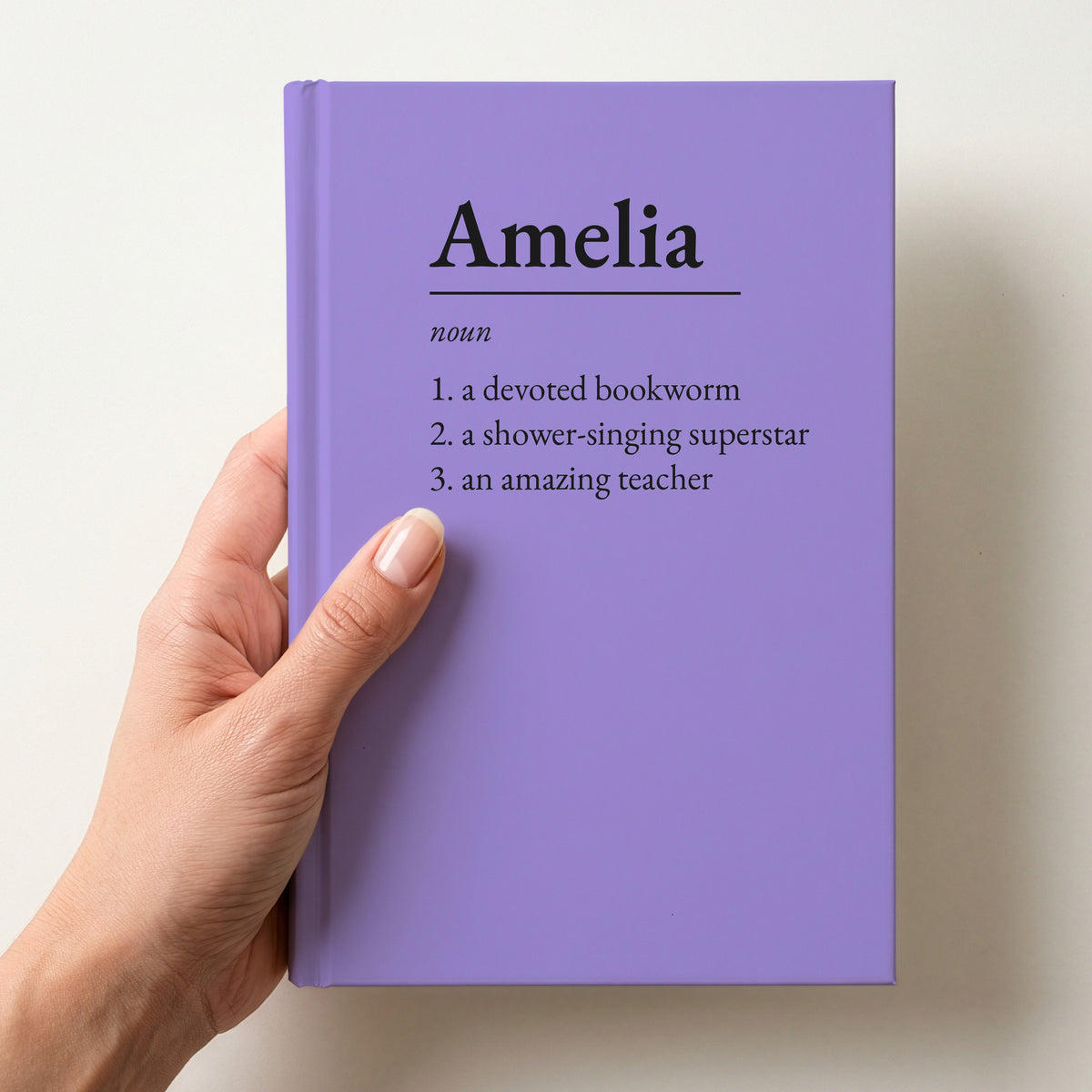 Hand holding a lilac personalised notebook with a dictionary definition of Amelia: a devoted bookworm, a shower-singing superstar, an amazing teacher