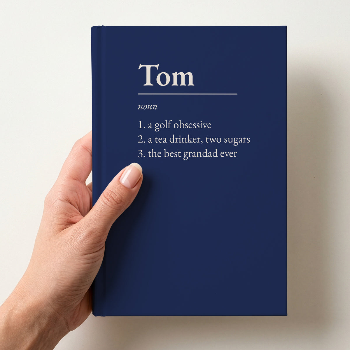 Hand holding a navy personalised notebook with a dictionary definition of Tom: a golf obsessive, a tea drinker, two sugars, the best grandad ever