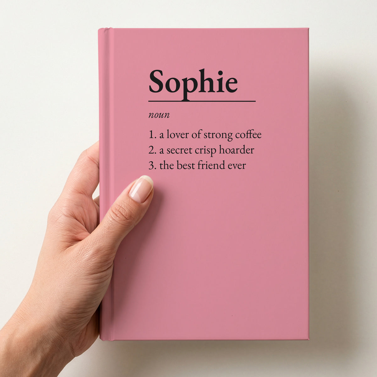 Hand holding a pink personalised notebook with a dictionary definition of Sophie: a lover of strong coffee, a secret crisp hoarder, the best friend ever
