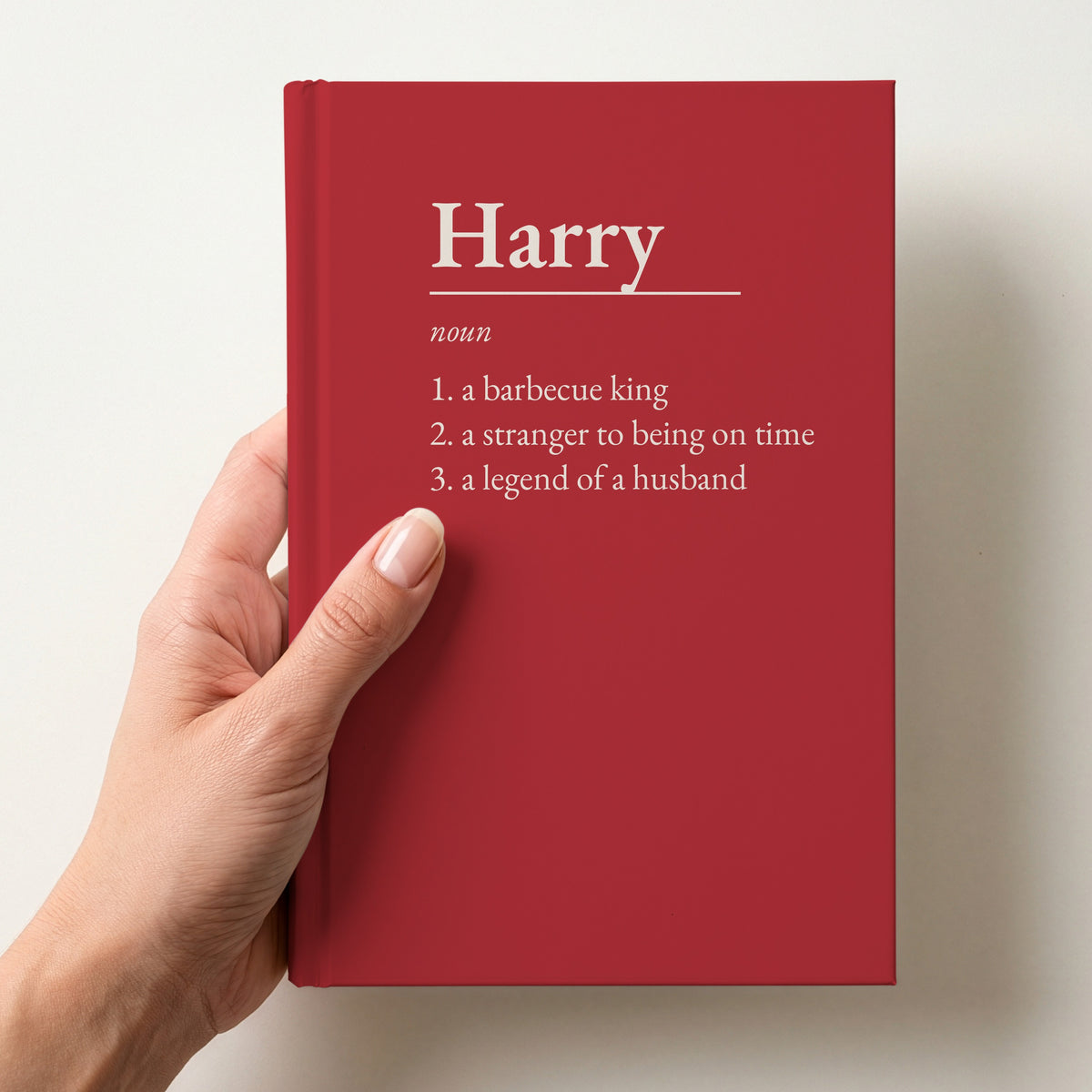 Hand holding a red personalised notebook with a dictionary definition of Harry: a barbecue king, a stranger to being on time, a legend of a husband