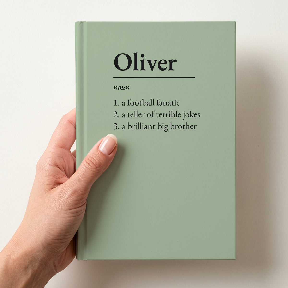Hand holding a sage personalised notebook with a dictionary definition of Oliver: a football fanatic, a teller of terrible jokes, a brilliant big brother