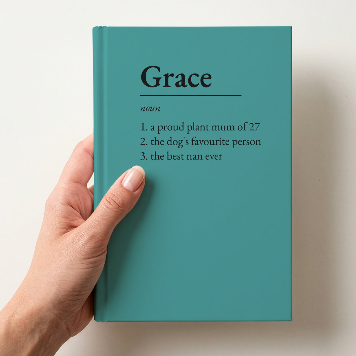 Hand holding a teal personalised notebook with a dictionary definition of Grace: a proud plant mum of 27, the dog's favourite person, the best nan ever