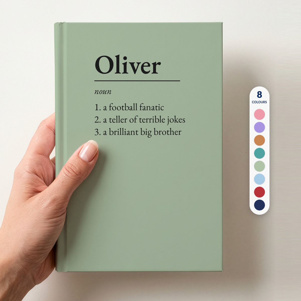 Hand holding a sage personalised notebook with a dictionary definition of Oliver: a football fanatic, a teller of terrible jokes, a brilliant big brother, beside a column of 8 colour swatches showing all 8 colourways