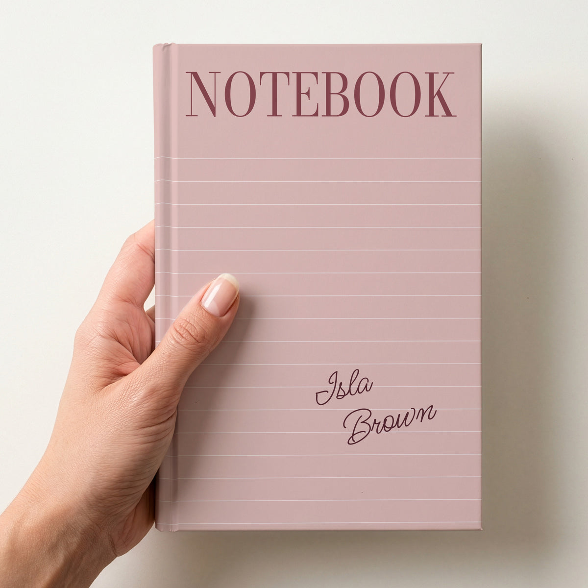 Hand holding a blush personalised notebook with a tall serif NOTEBOOK title, ruled lines and the name Isla Brown in handwritten script