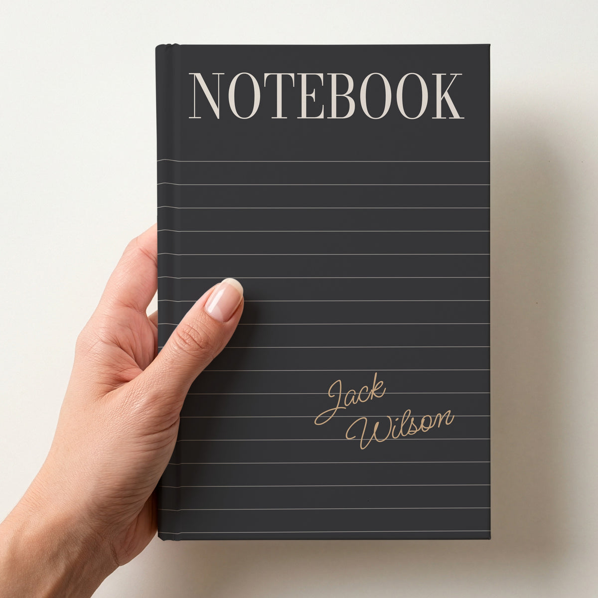 Hand holding a charcoal personalised notebook with a tall serif NOTEBOOK title, ruled lines and the name Jack Wilson in handwritten script
