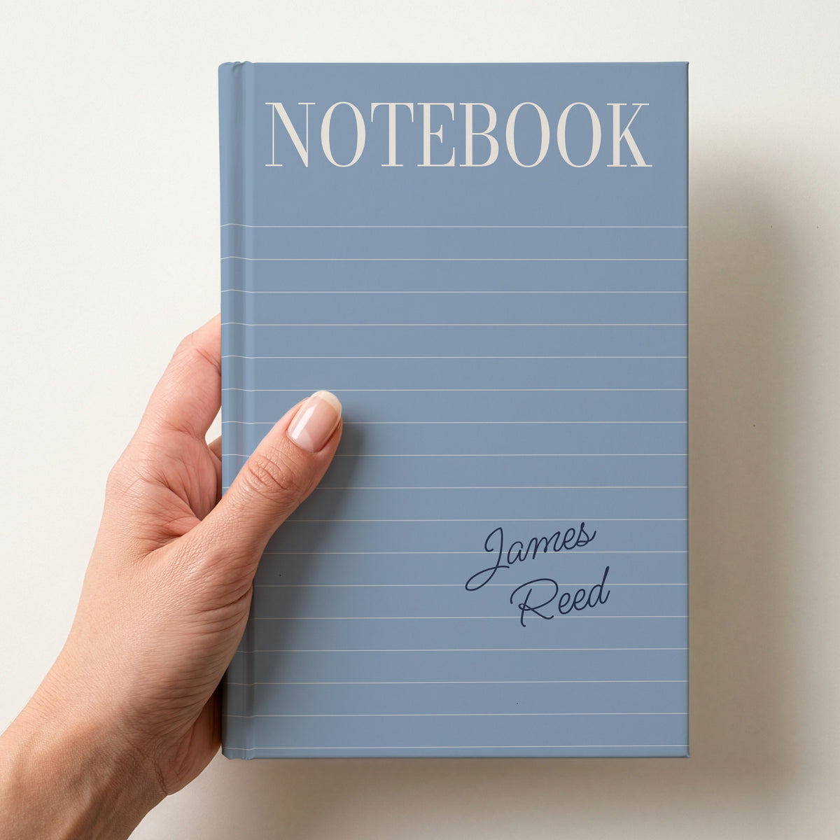 Hand holding a dusty blue personalised notebook with a tall serif NOTEBOOK title, ruled lines and the name James Reed in handwritten script