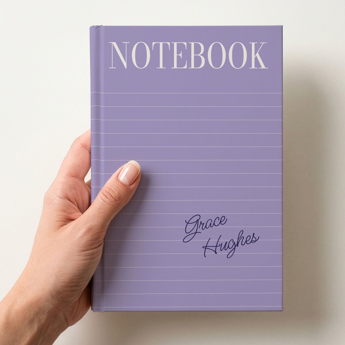 Hand holding a lilac personalised notebook with a tall serif NOTEBOOK title, ruled lines and the name Grace Hughes in handwritten script