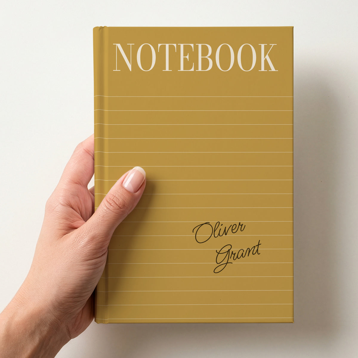 Hand holding a mustard personalised notebook with a tall serif NOTEBOOK title, ruled lines and the name Oliver Grant in handwritten script