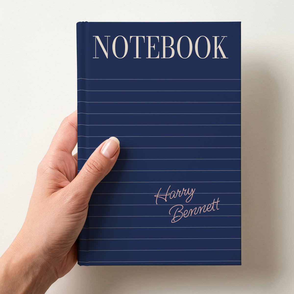 Hand holding a navy personalised notebook with a tall serif NOTEBOOK title, ruled lines and the name Harry Bennett in handwritten script