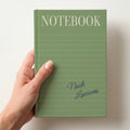 Hand holding a sage personalised notebook with a tall serif NOTEBOOK title, ruled lines and the name Noah Luciano in handwritten script