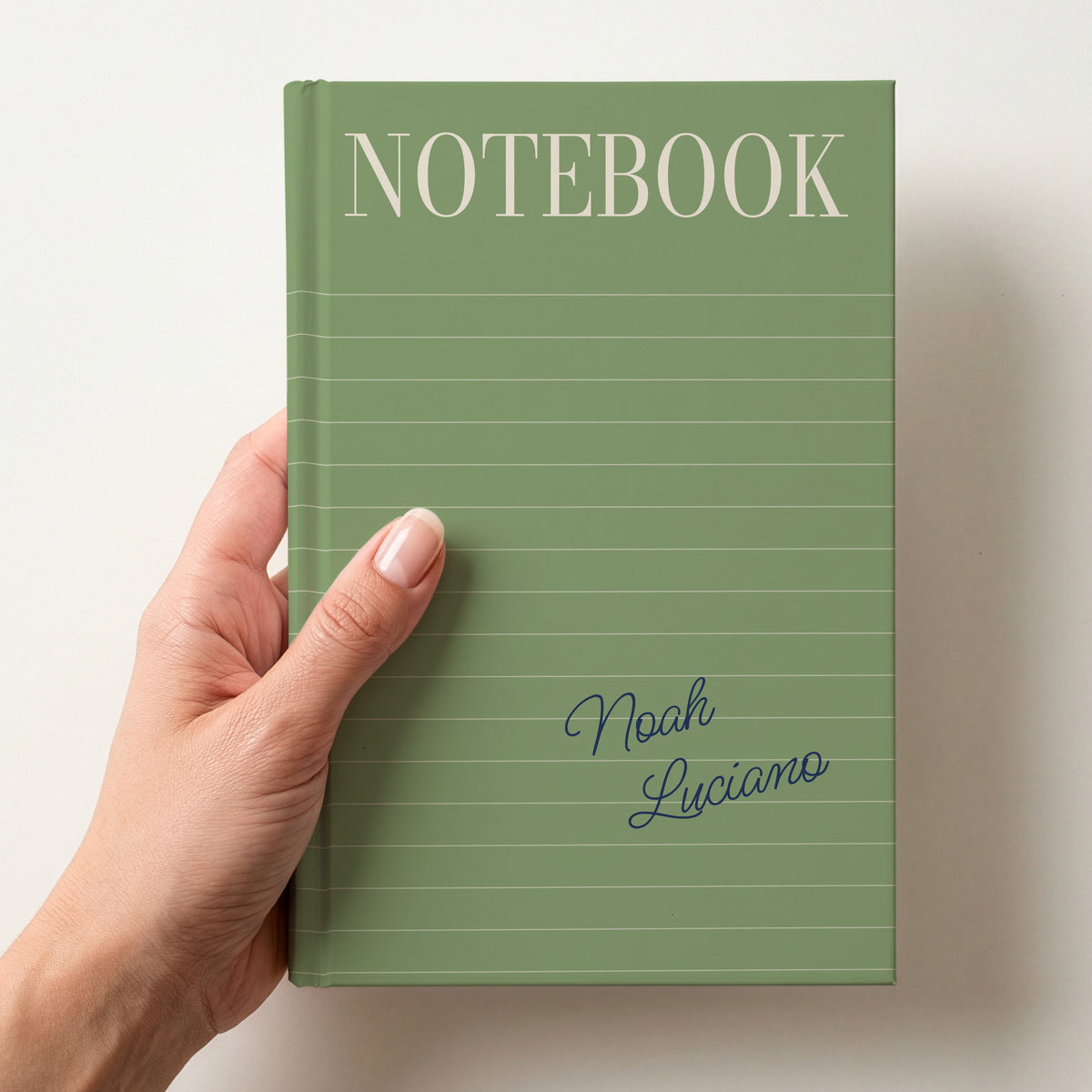 Hand holding a sage personalised notebook with a tall serif NOTEBOOK title, ruled lines and the name Noah Luciano in handwritten script