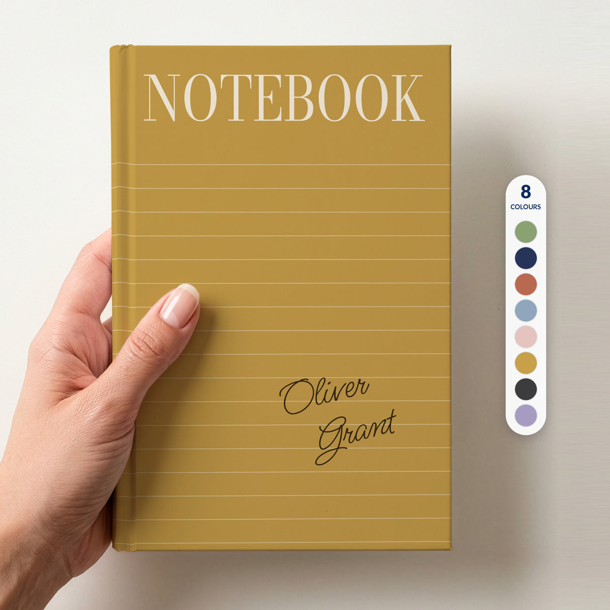 Hand holding a mustard personalised notebook with a tall serif NOTEBOOK title, ruled lines and the name Oliver Grant in handwritten script, beside a column of 8 colour swatches showing all 8 colourways