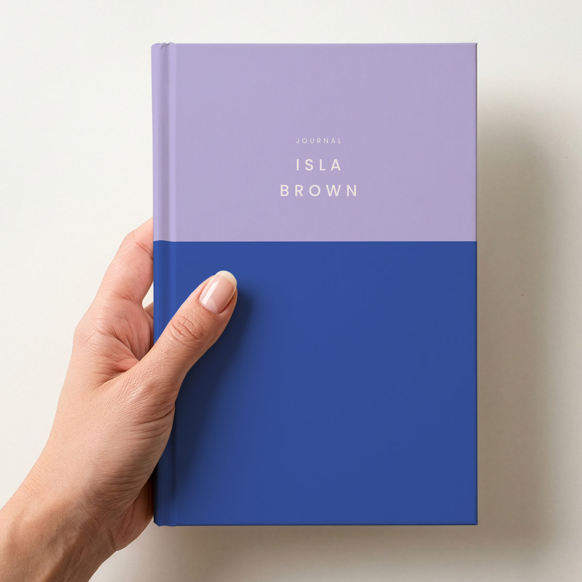 Hand holding a lilac & cobalt colour block notebook with JOURNAL and the name Isla Brown in spaced capitals