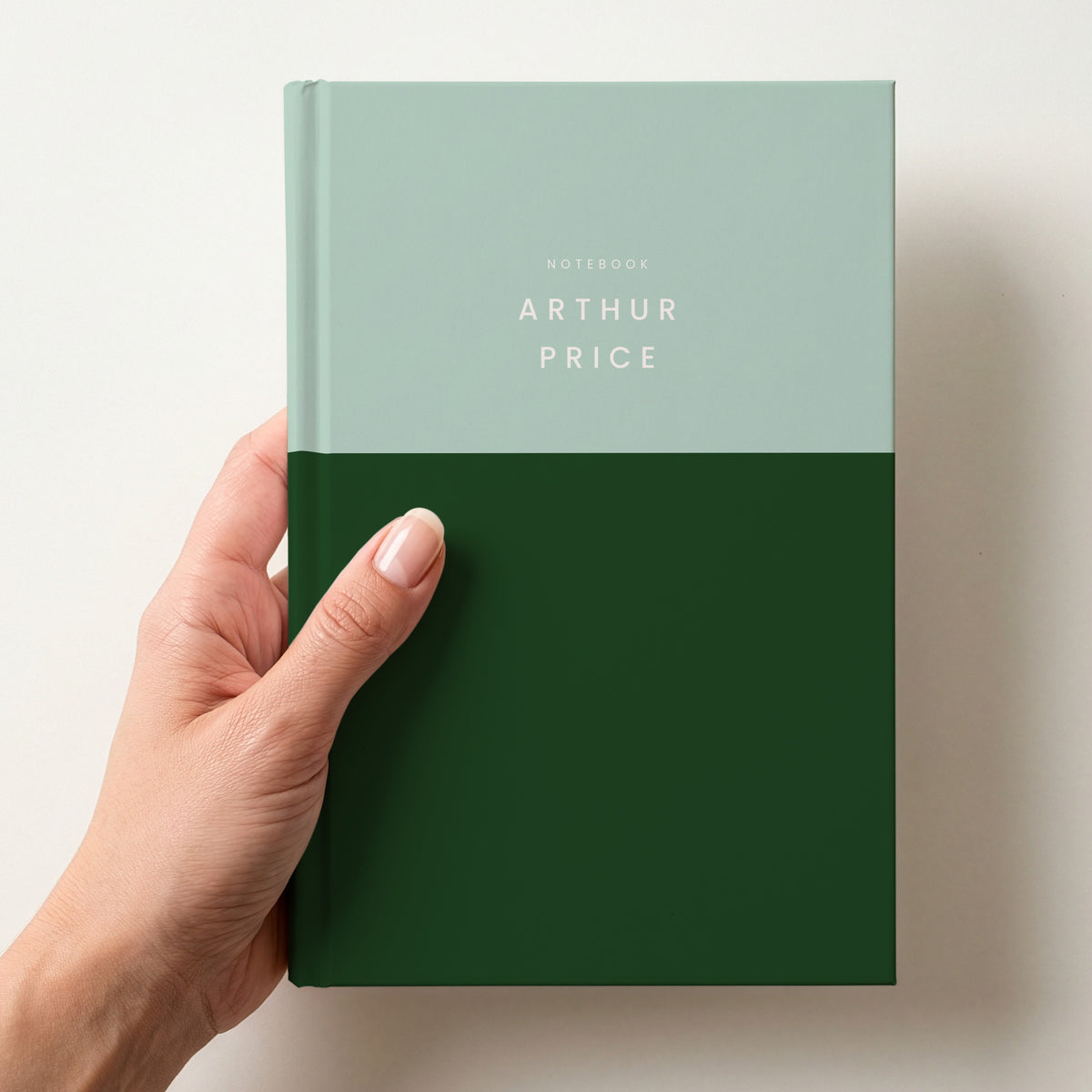 Hand holding a mint & bottle colour block notebook with NOTEBOOK and the name Arthur Price in spaced capitals