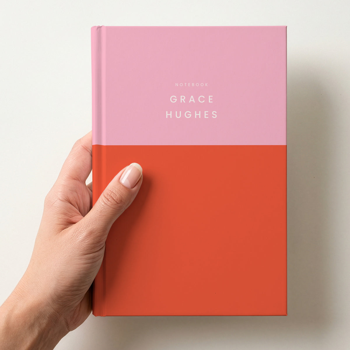 Hand holding a pink & red colour block notebook with NOTEBOOK and the name Grace Hughes in spaced capitals