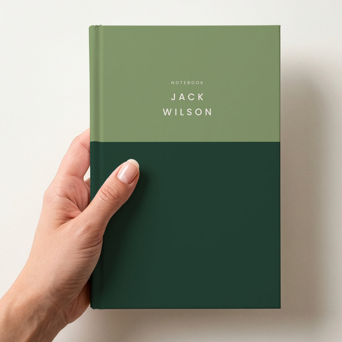 Hand holding a sage & pine colour block notebook with NOTEBOOK and the name Jack Wilson in spaced capitals