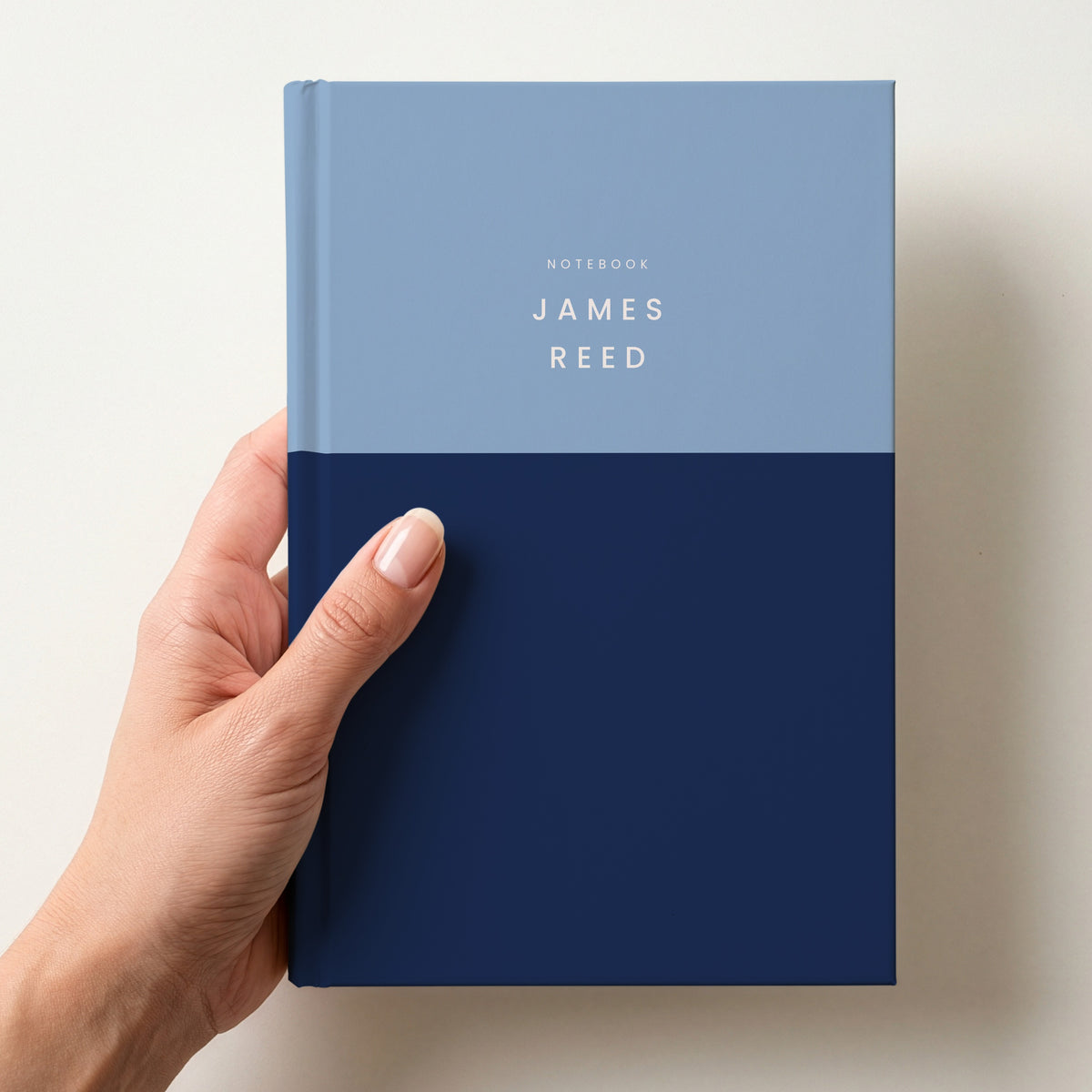 Hand holding a sky & navy colour block notebook with NOTEBOOK and the name James Reed in spaced capitals