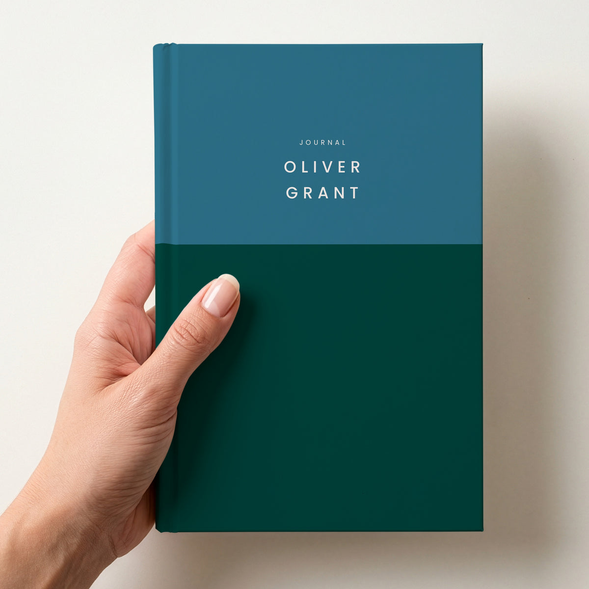 Hand holding a teal & forest colour block notebook with JOURNAL and the name Oliver Grant in spaced capitals