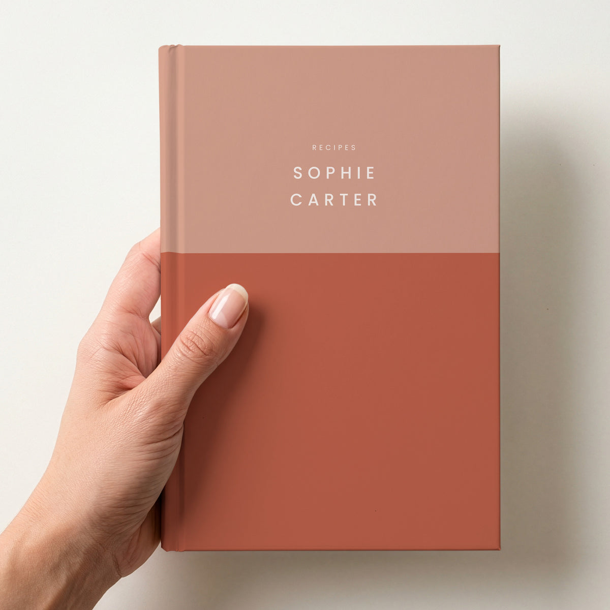 Hand holding a terracotta colour block notebook with RECIPES and the name Sophie Carter in spaced capitals