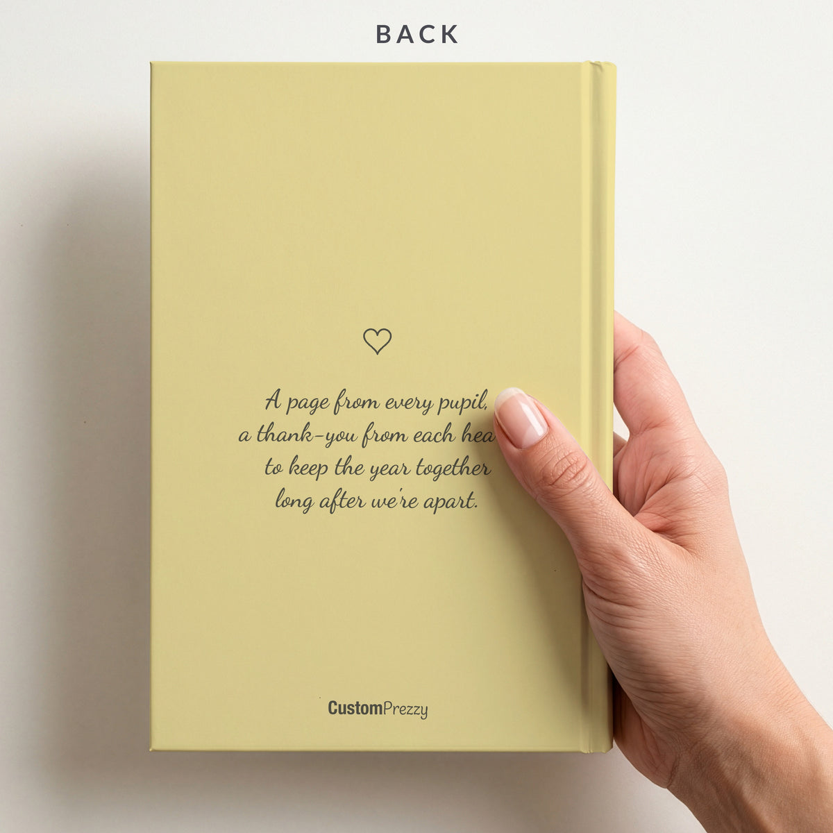 Back of the butter yellow autograph book held in a hand, with a small heart, the verse A page from every pupil, a thank-you from each heart, and the Custom Prezzy wordmark