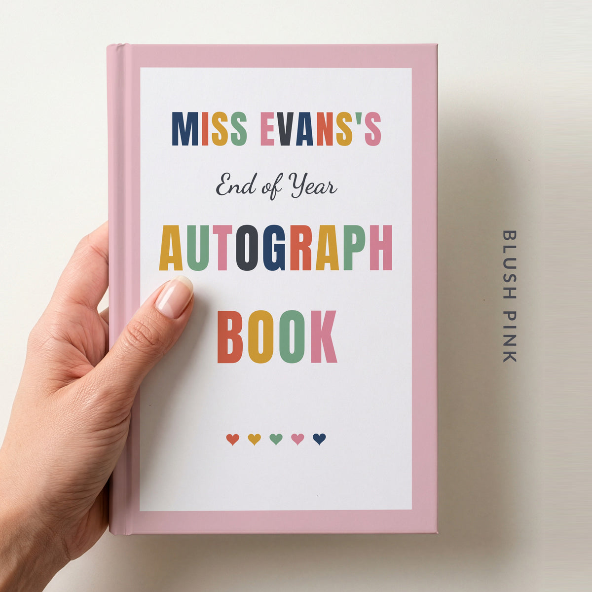Hand holding a personalised teacher autograph book with a blush pink border, reading MISS EVANS'S, End of Year AUTOGRAPH BOOK and a row of colourful hearts