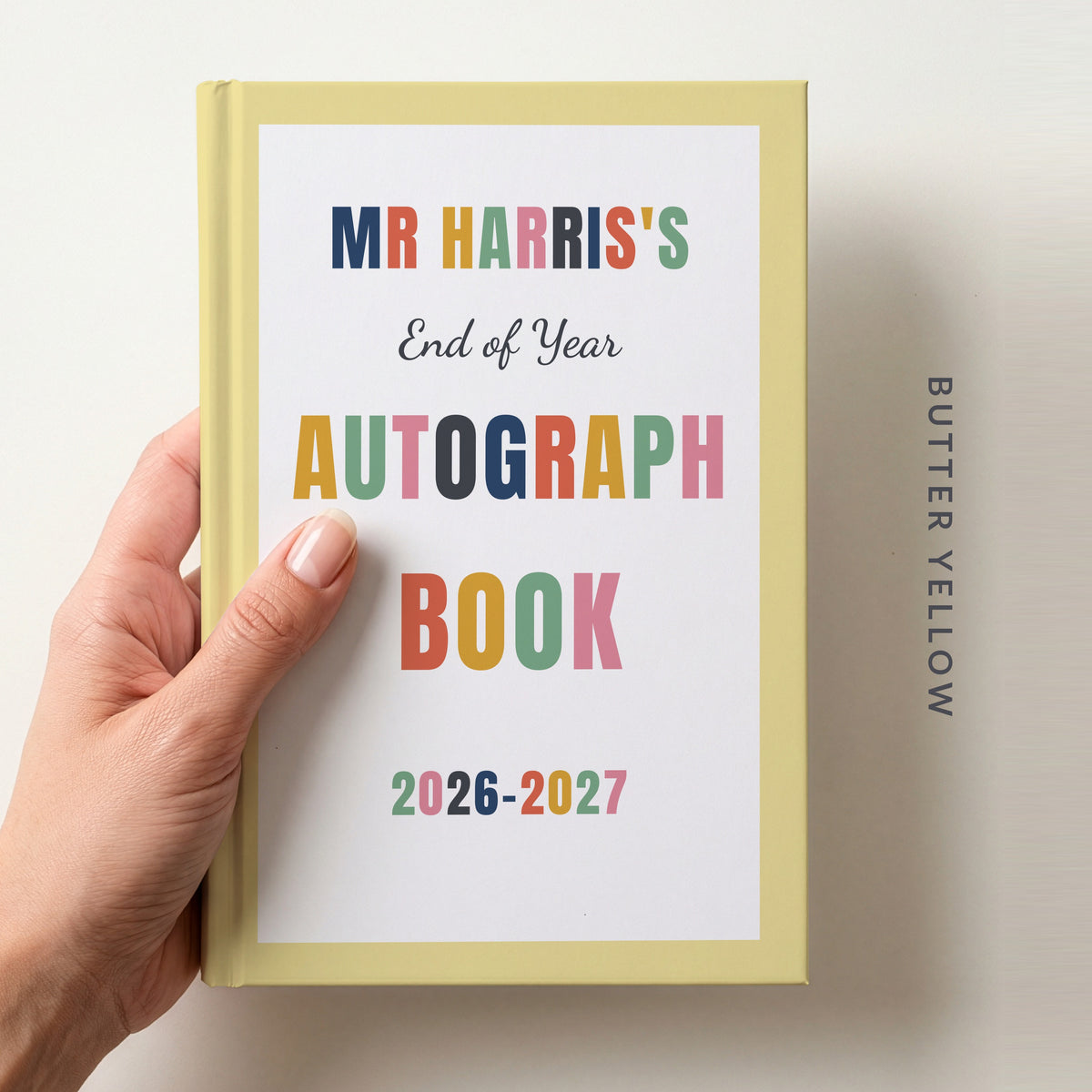 Hand holding a personalised teacher autograph book with a butter yellow border, reading MR HARRIS'S, End of Year AUTOGRAPH BOOK and 2026–2027