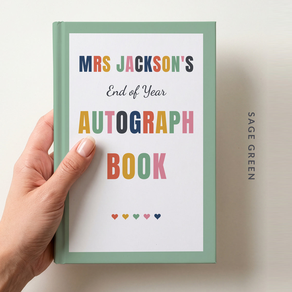 Hand holding a personalised teacher autograph book with a sage green border, reading MRS JACKSON'S, End of Year AUTOGRAPH BOOK and a row of colourful hearts