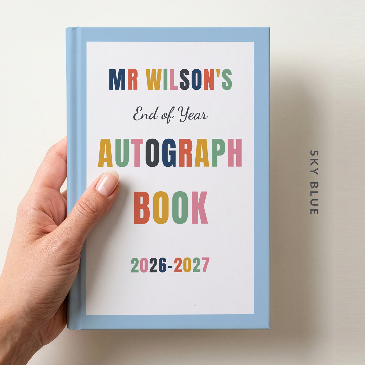 Hand holding a personalised teacher autograph book with a sky blue border, reading MR WILSON'S, End of Year AUTOGRAPH BOOK and 2026–2027
