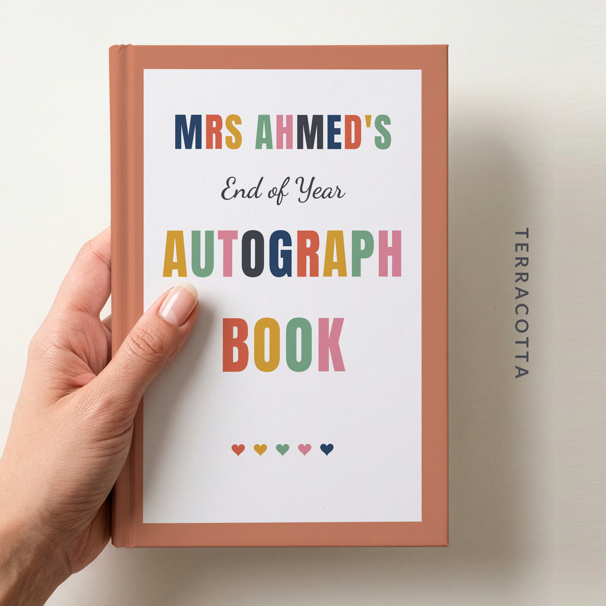 Hand holding a personalised teacher autograph book with a terracotta border, reading MRS AHMED'S, End of Year AUTOGRAPH BOOK and a row of colourful hearts