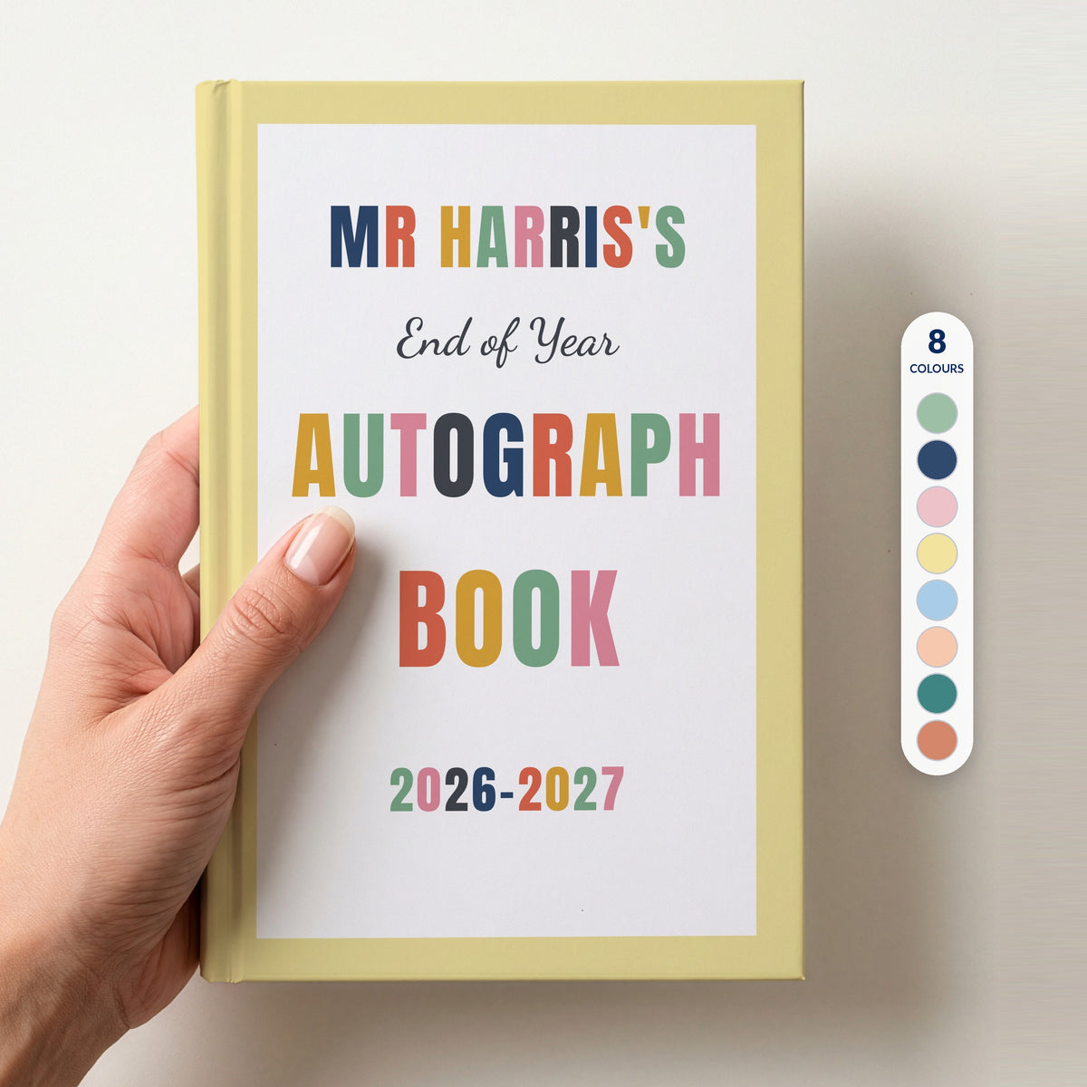 Hand holding a personalised teacher autograph book with a butter yellow border, reading MR HARRIS'S, End of Year AUTOGRAPH BOOK and 2026–2027, beside a column of 8 colour swatches showing all 8 border colours