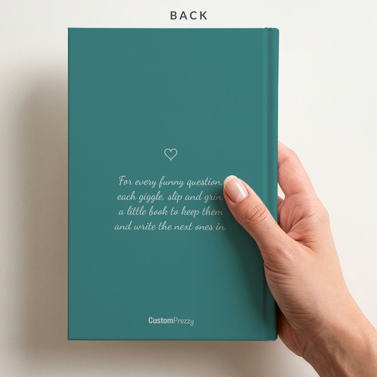 Back of the teal journal held in a hand, with a small heart, the verse For every funny question, each giggle, slip and grin, and the Custom Prezzy wordmark