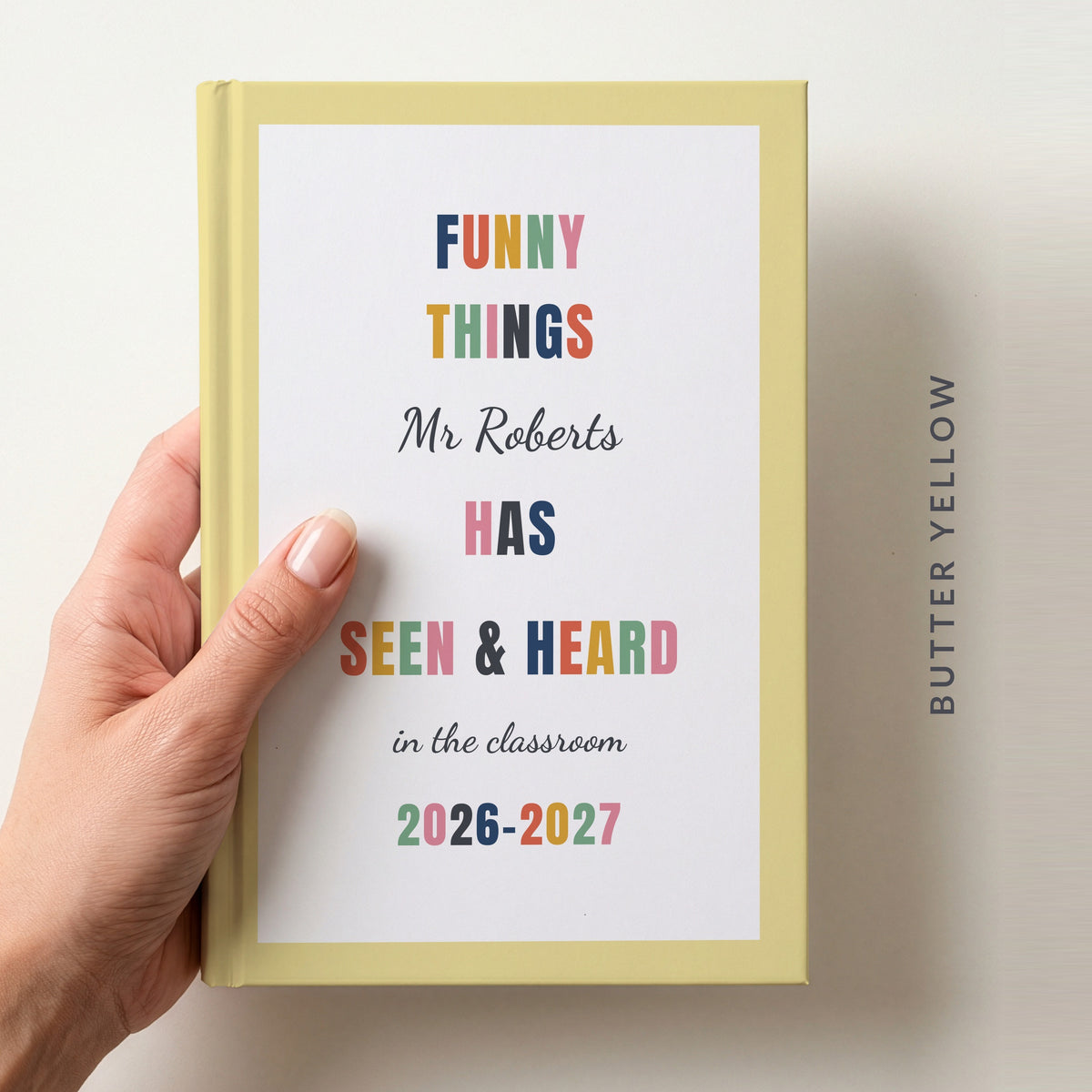 Hand holding a personalised teacher journal with a butter yellow border, reading FUNNY THINGS Mr Roberts HAS SEEN & HEARD in the classroom, and 2026–2027