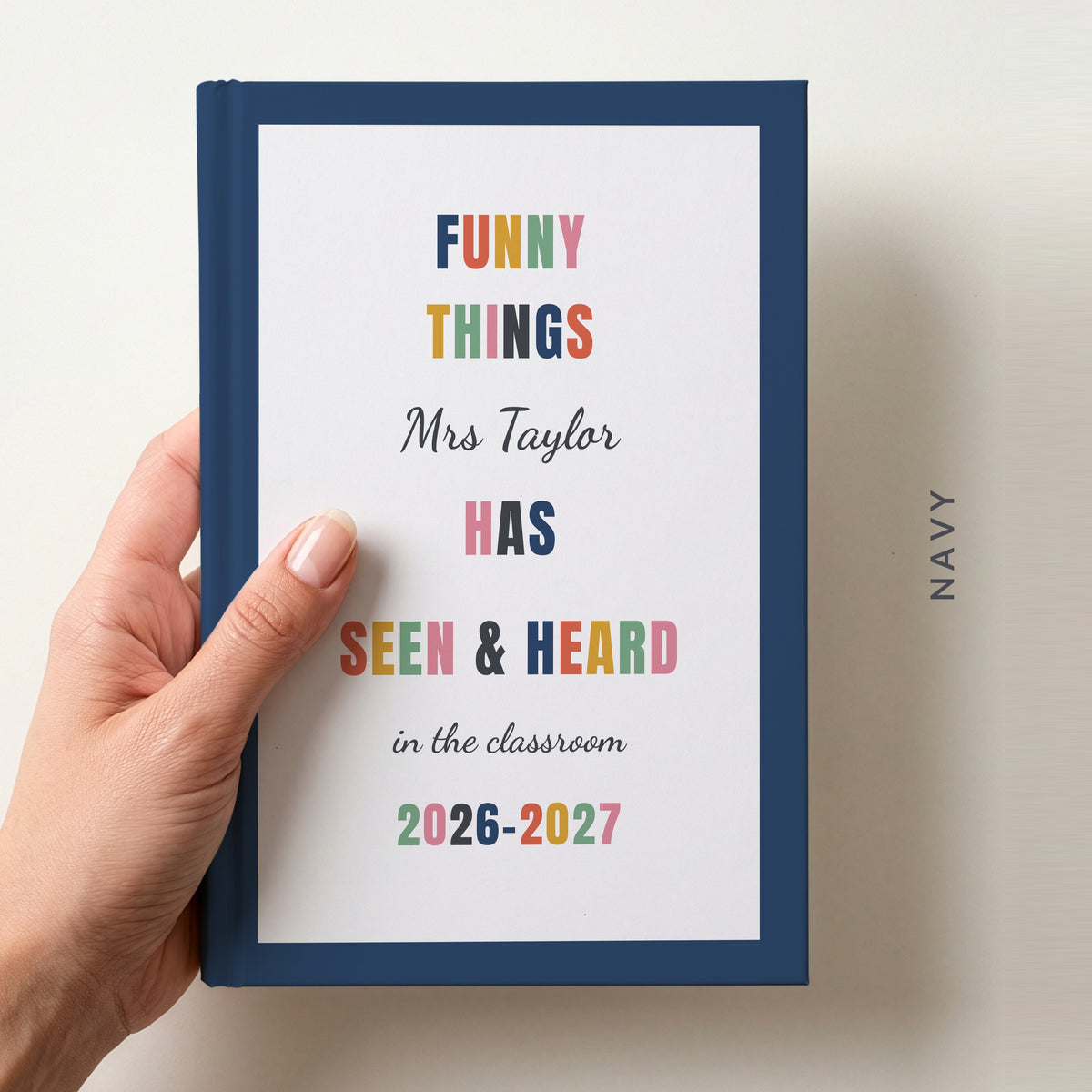 Hand holding a personalised teacher journal with a navy border, reading FUNNY THINGS Mrs Taylor HAS SEEN & HEARD in the classroom, and 2026–2027