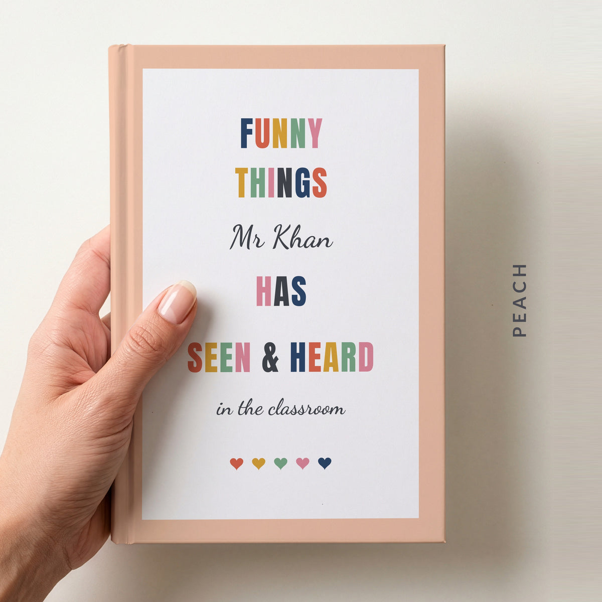 Hand holding a personalised teacher journal with a peach border, reading FUNNY THINGS Mr Khan HAS SEEN & HEARD in the classroom, and a row of colourful hearts
