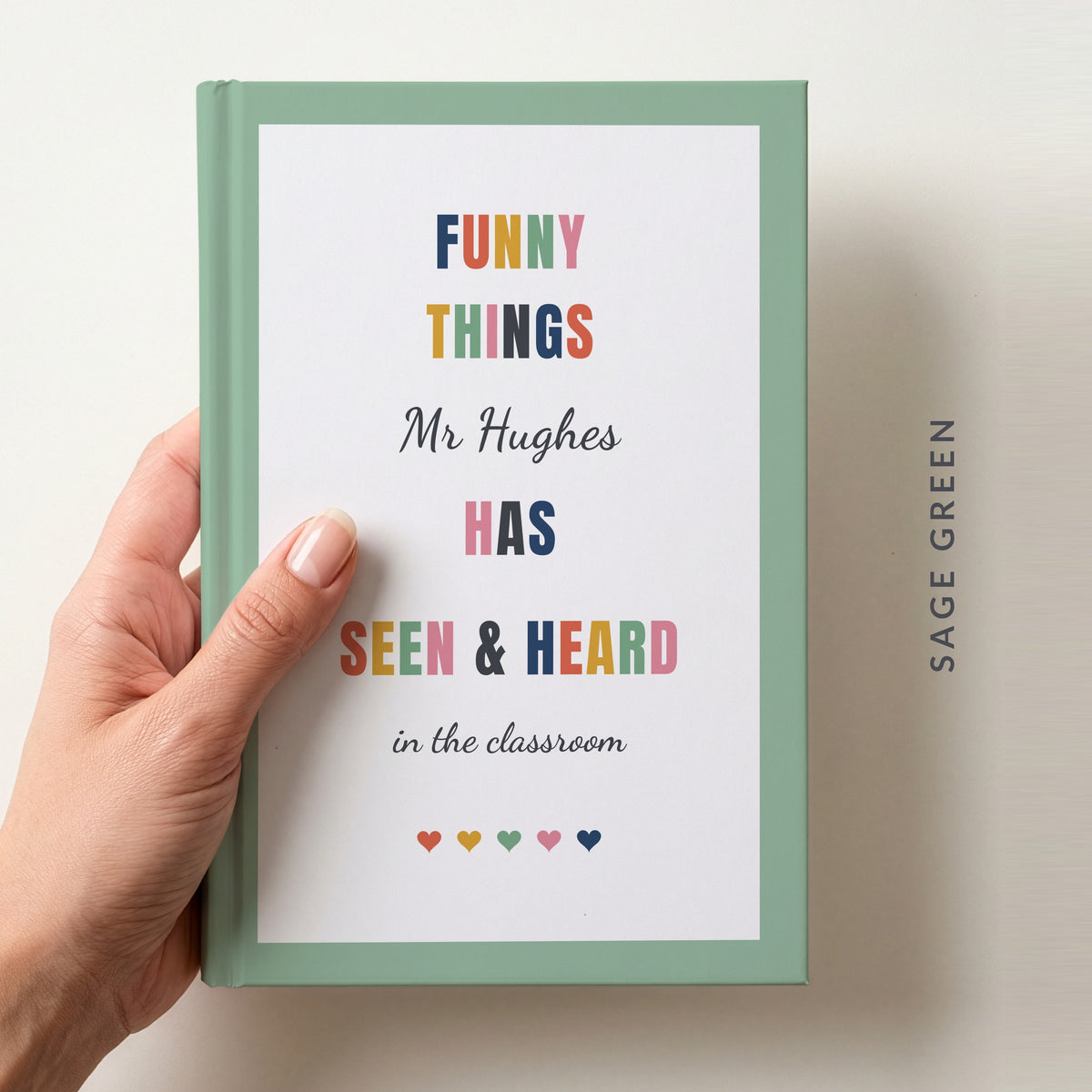 Hand holding a personalised teacher journal with a sage green border, reading FUNNY THINGS Mr Hughes HAS SEEN & HEARD in the classroom, and a row of colourful hearts