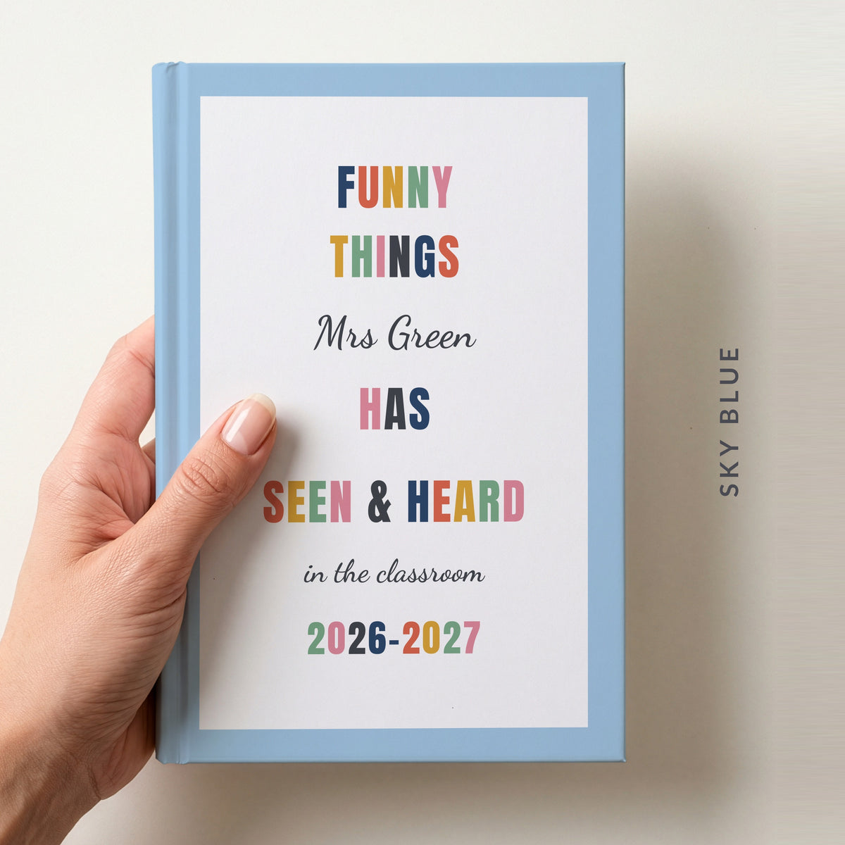 Hand holding a personalised teacher journal with a sky blue border, reading FUNNY THINGS Mrs Green HAS SEEN & HEARD in the classroom, and 2026–2027