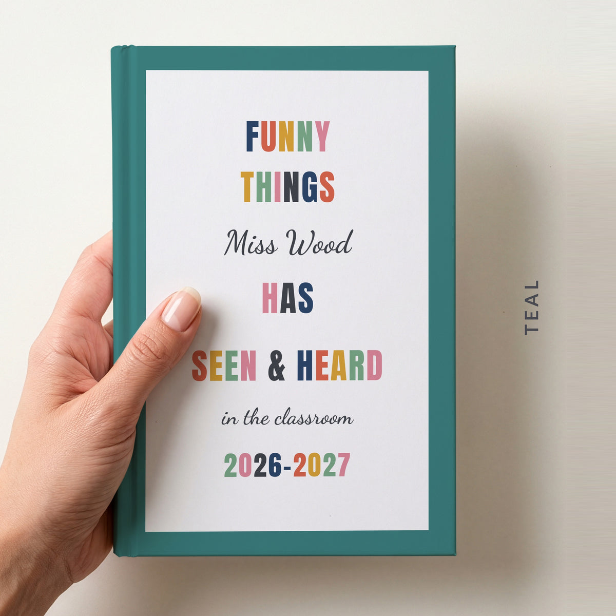 Hand holding a personalised teacher journal with a teal border, reading FUNNY THINGS Miss Wood HAS SEEN & HEARD in the classroom, and 2026–2027