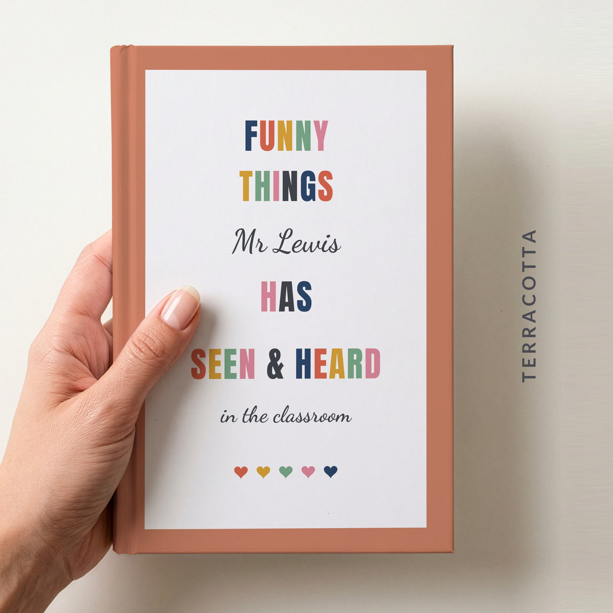 Hand holding a personalised teacher journal with a terracotta border, reading FUNNY THINGS Mr Lewis HAS SEEN & HEARD in the classroom, and a row of colourful hearts