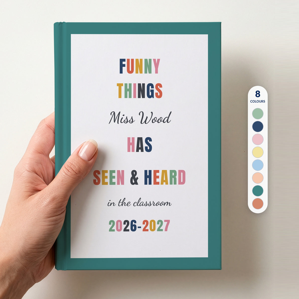 Hand holding a personalised teacher journal with a teal border, reading FUNNY THINGS Miss Wood HAS SEEN & HEARD in the classroom, and 2026–2027, beside a column of 8 colour swatches showing all 8 border colours