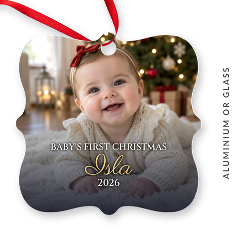 Personalised Baby's First Christmas photo ornament for Isla, 2026: a scalloped aluminium ornament printed with a photo of a smiling baby girl by the Christmas tree, available in aluminium or glass